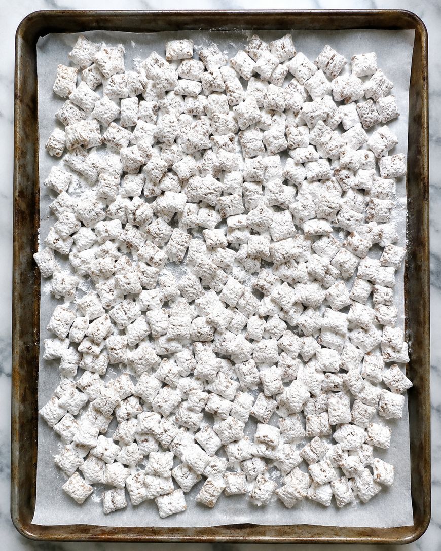 A baking sheet lined with parchment paper is covered with Chex cereal coated in powdered sugar, resembling homemade puppy chow or Muddy Buddies.