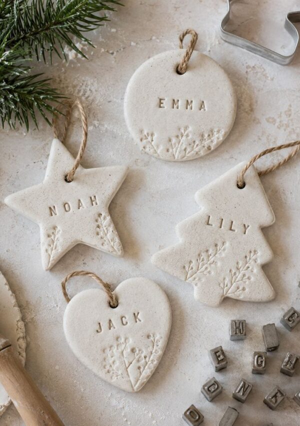 Four white clay ornaments—star, circle, heart, and tree—each stamped with a different name (Noah, Emma, Jack, Lily) and decorated with floral imprints. Crafting tools and letter stamps are nearby.