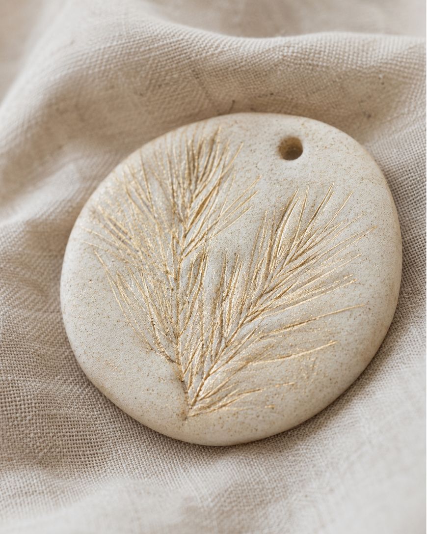 Round beige clay ornament with a hole at the top, featuring a delicate gold-etched botanical or pine needle design, resting on a soft, textured fabric background.