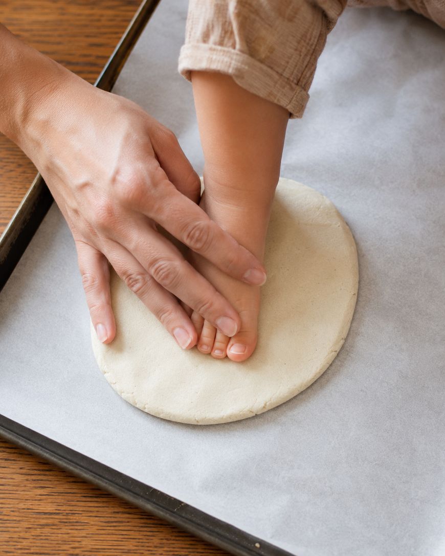 An adult presses a childs bare foot into a round piece of dough on a parchment-lined baking tray, likely to make a footprint impression.