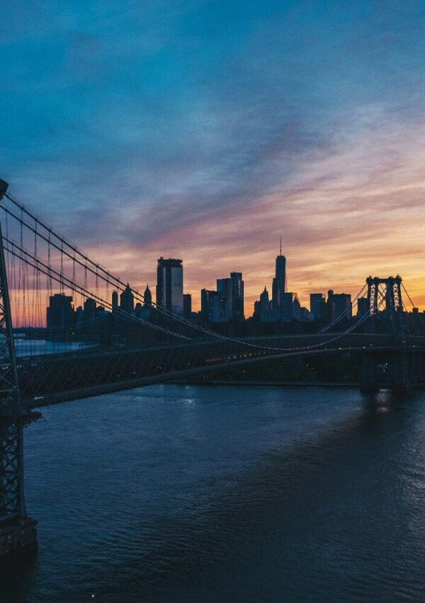 A city skyline at sunset with a suspension bridge crossing over a river, silhouetted against a colorful, cloudy sky.