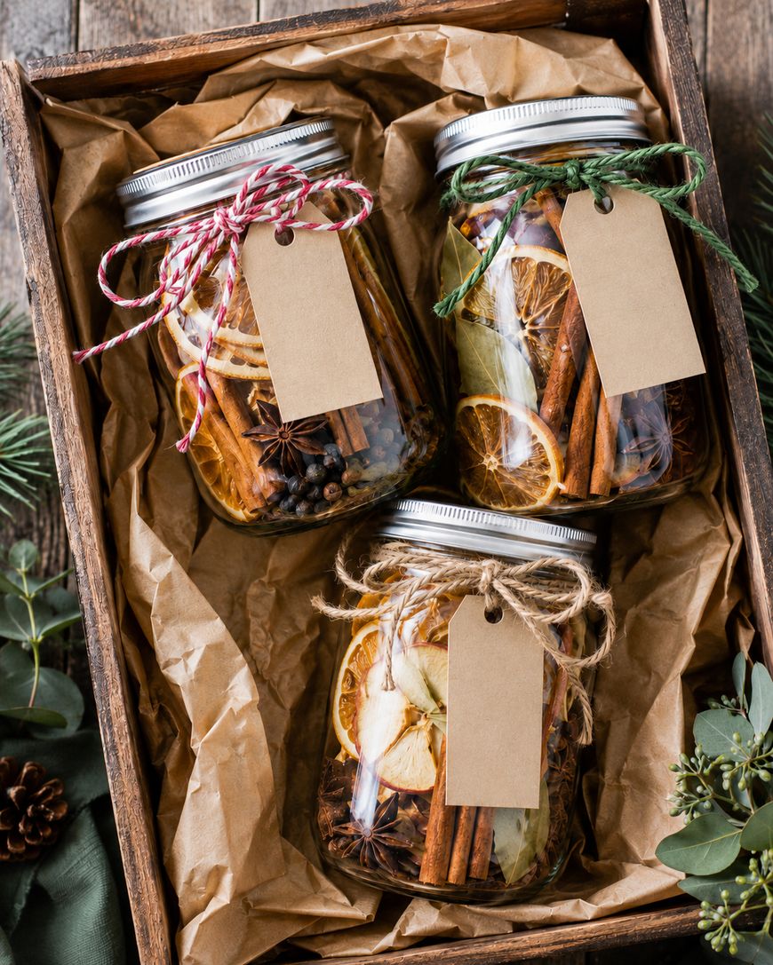 Three glass jars filled with dried citrus slices, cinnamon sticks, and spices, each topped with a blank brown tag and twine, are arranged in a wooden box lined with crumpled brown paper.