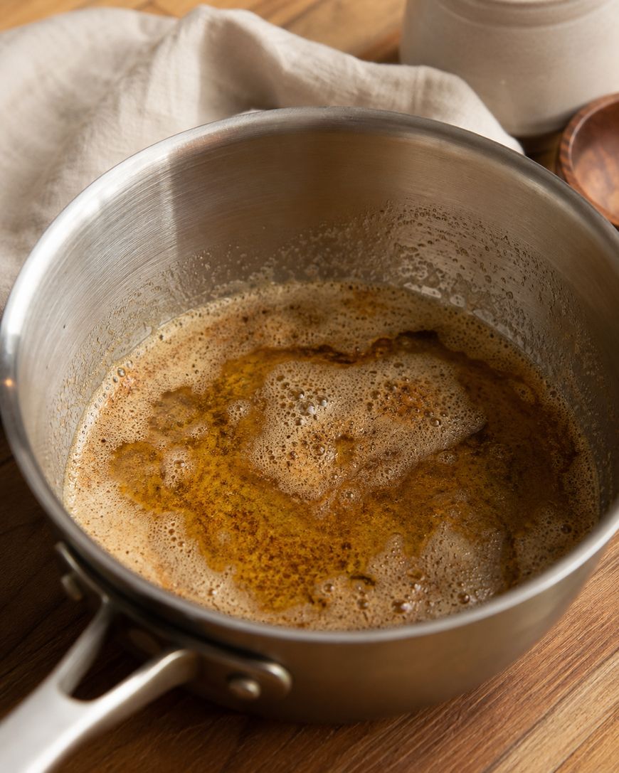 A silver saucepan contains bubbling brown butter, sitting on a wooden countertop with a beige cloth and a small wooden dish nearby.