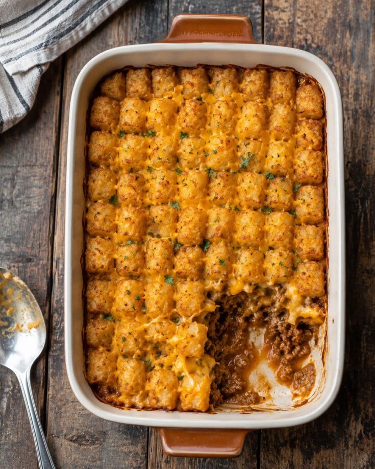 A baked casserole topped with golden tater tots, some removed to reveal a layer of ground beef and melted cheese underneath, sits on a wooden table beside a spoon and a striped cloth.