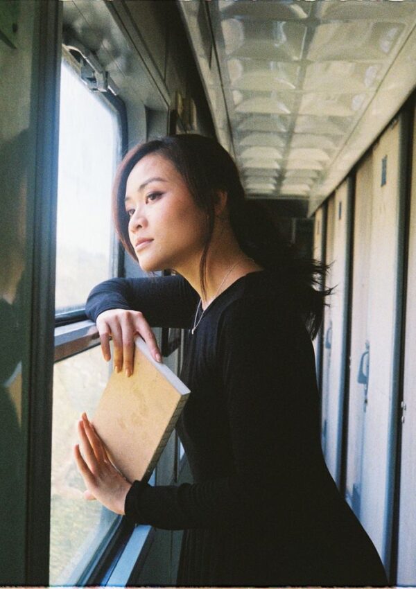A woman in a black dress stands in a train corridor, holding a closed book and looking thoughtfully out the window.