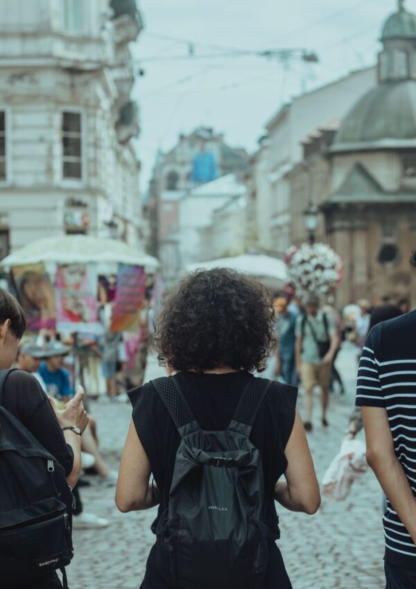 Three people with backpacks walk down a busy cobblestone street lined with historic buildings and market stalls, seen from behind. The street is lively with pedestrians and street vendors.