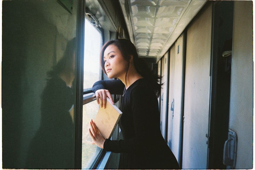 A woman in a black dress holds a book and gazes thoughtfully out the window of a train, sunlight softly illuminating her face.