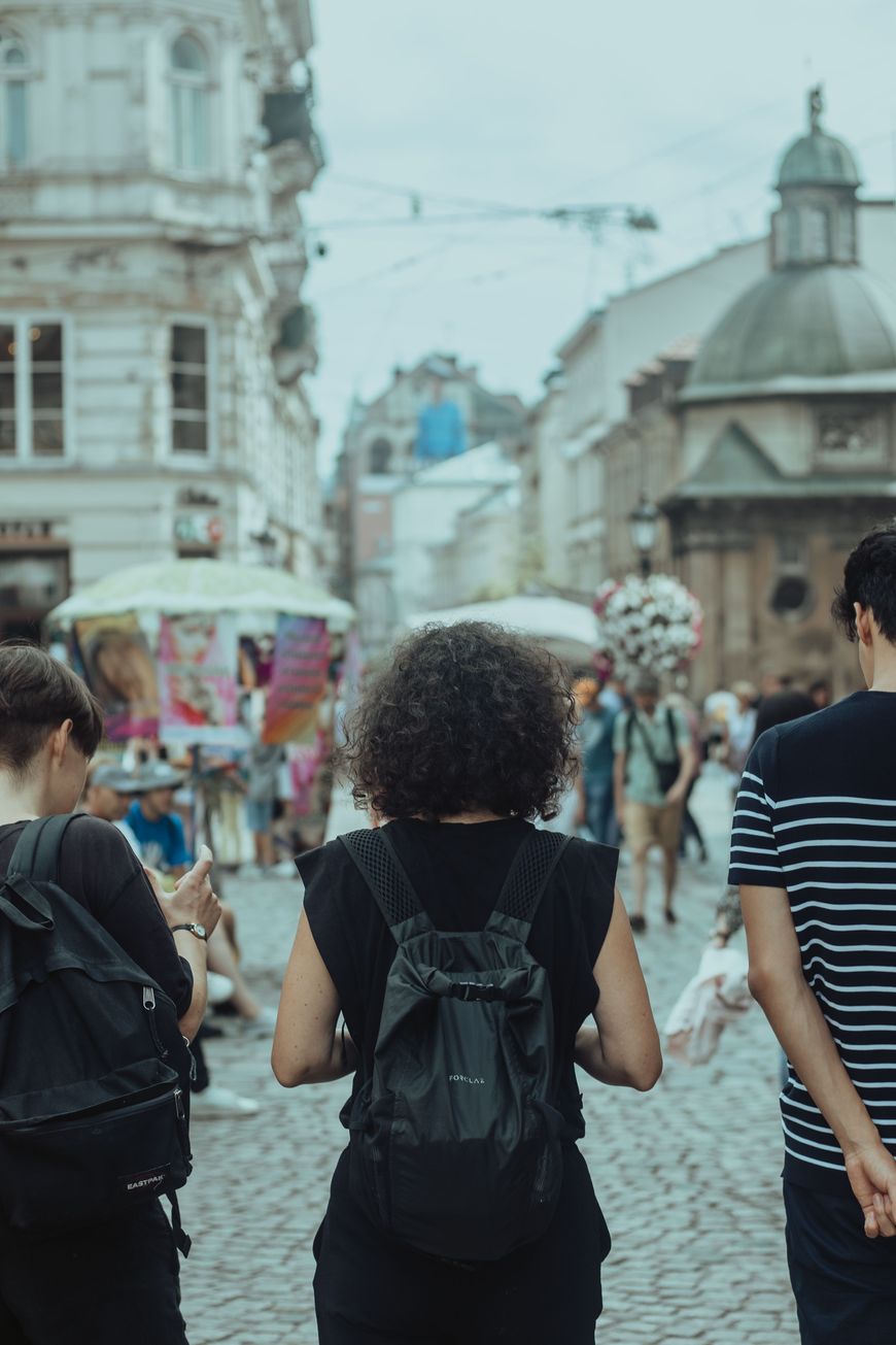 Three people with backpacks walk on a cobblestone street in a city, surrounded by historic buildings and market stalls. The scene is lively, with other pedestrians visible in the background.