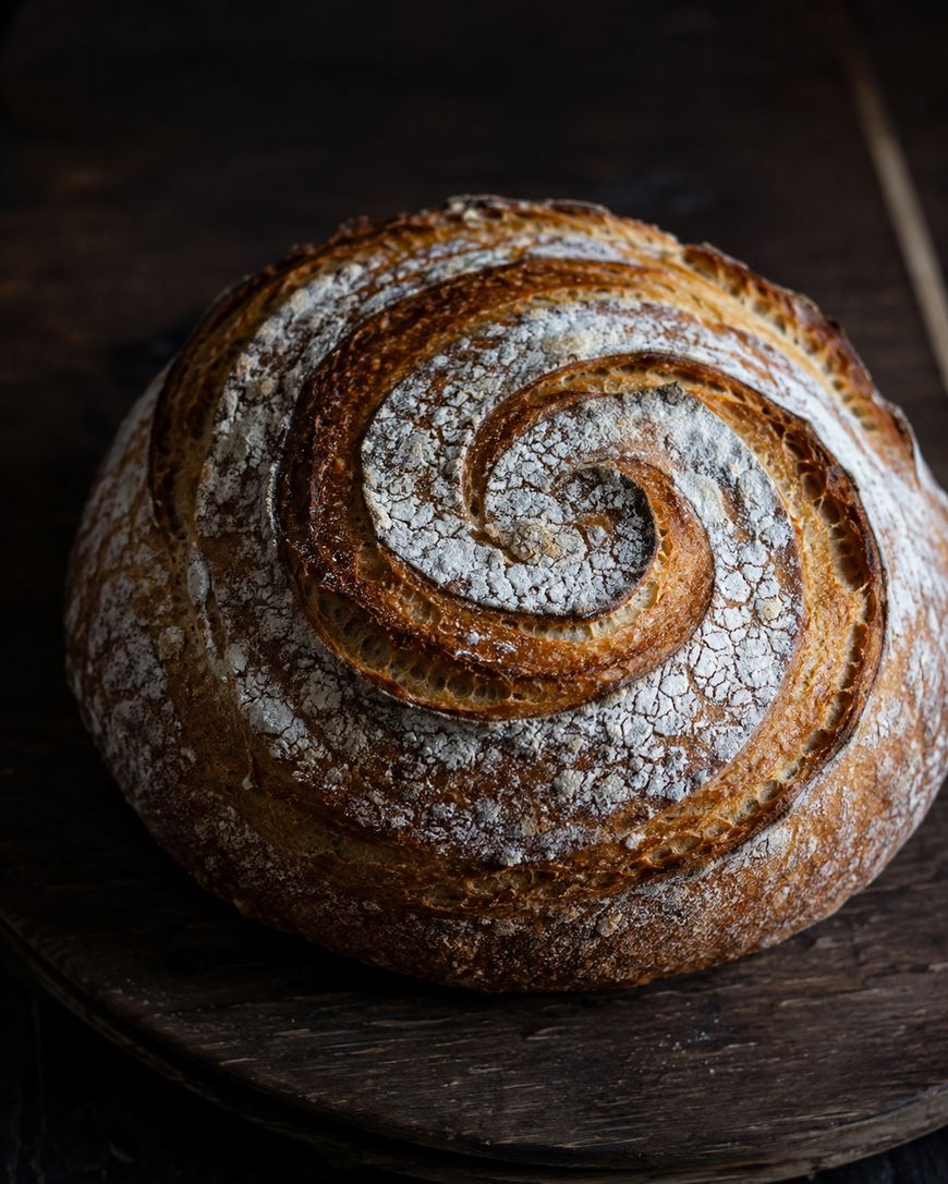 A round loaf of artisan bread with a golden-brown crust and a spiral pattern of flour and scoring on top, set on a dark wooden surface.