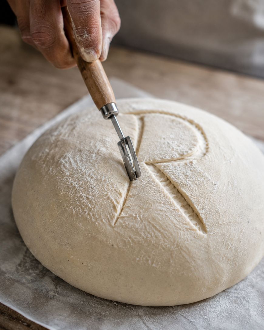 A hand uses a bread lame to carve the letter R into a round, floured dough loaf on parchment paper, preparing it for baking.