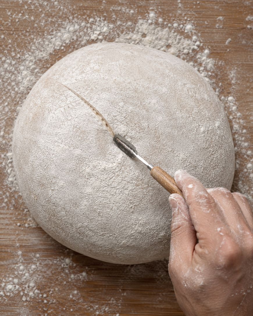 A hand uses a bread lame to score a line on a round, floured dough ball on a wooden surface dusted with flour.