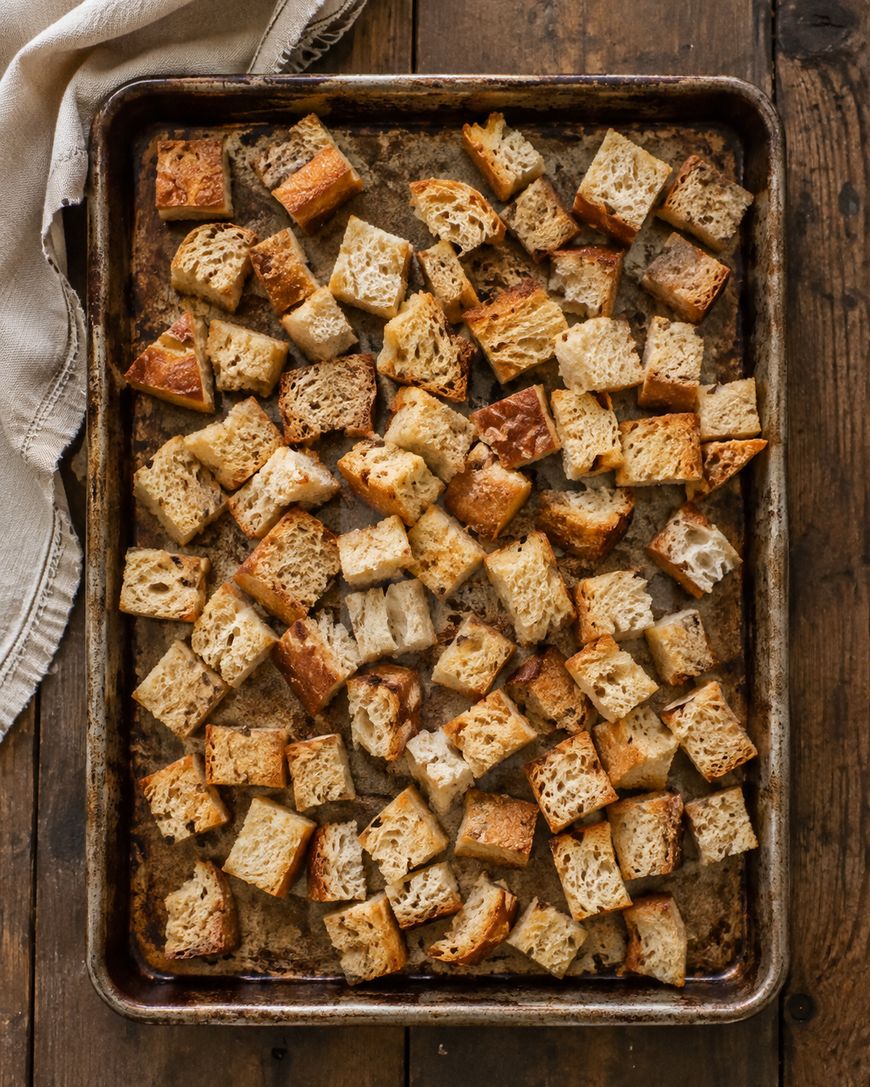 A baking sheet with evenly spread, golden-brown bread cubes sits on a rustic wooden table, next to a folded beige cloth.