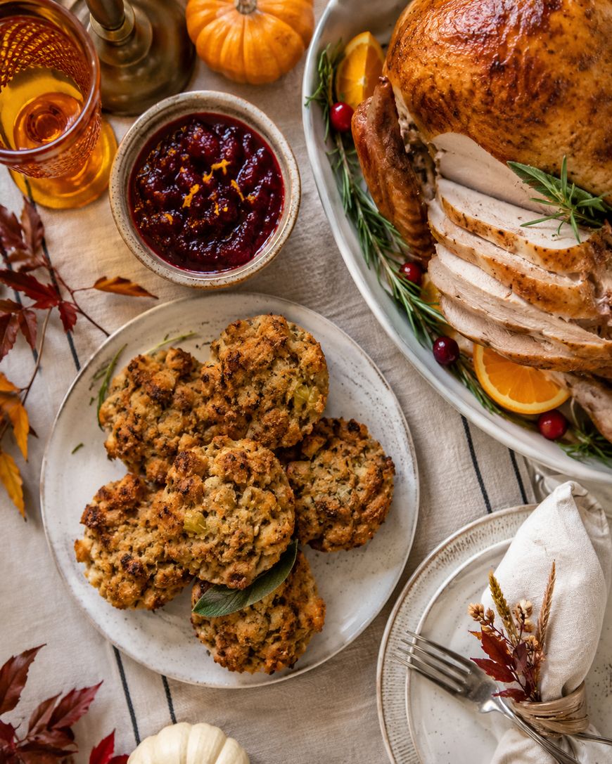 A Thanksgiving table with sliced roast turkey, a plate of stuffing, cranberry sauce, festive drinks, and autumn leaves on a cream tablecloth.