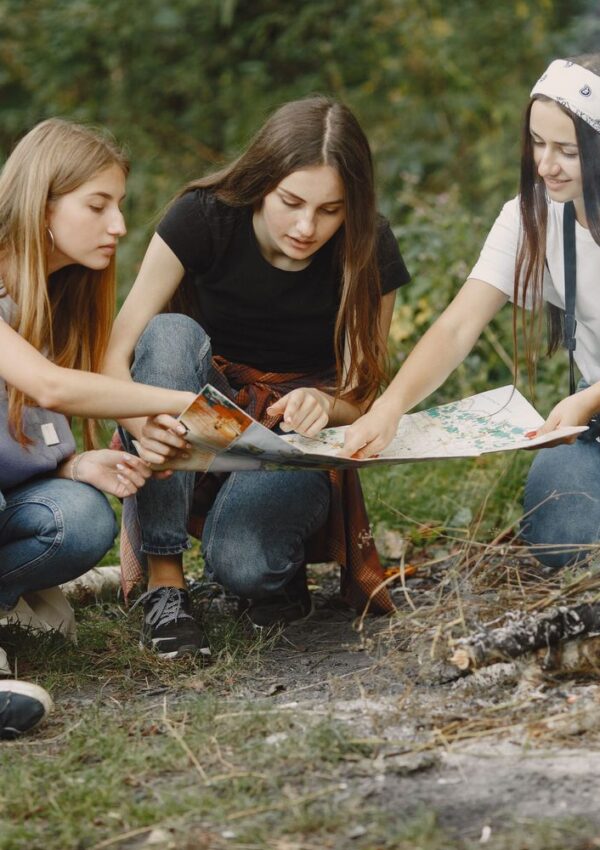 Three young women crouch near a campfire, closely examining a map together outdoors. Trees and greenery are visible in the background.