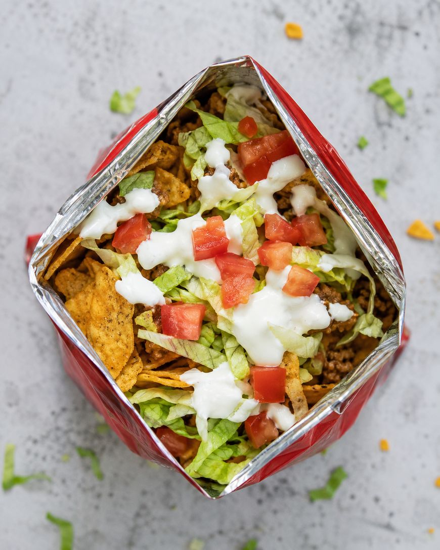 An open chip bag filled with nachos, ground meat, shredded lettuce, diced tomatoes, and a drizzle of white sauce, viewed from above on a light surface.