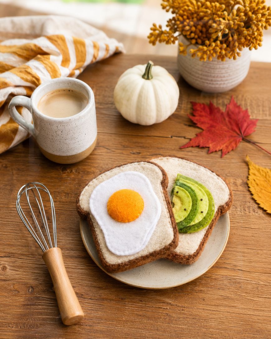 Plate with two slices of bread topped with felt egg and avocado, surrounded by a whisk, a mug, a white pumpkin, autumn leaves, and a vase of yellow flowers on a wooden table.