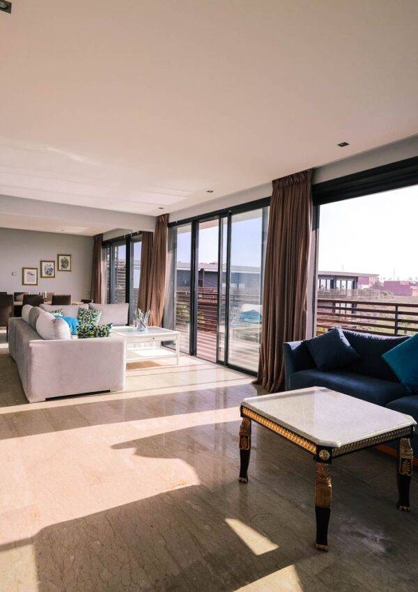 Modern living room with large windows, sunlight streaming in, two sofas with colorful cushions, a coffee table, and a balcony view outside. Marble flooring and neutral walls create a bright, spacious atmosphere.