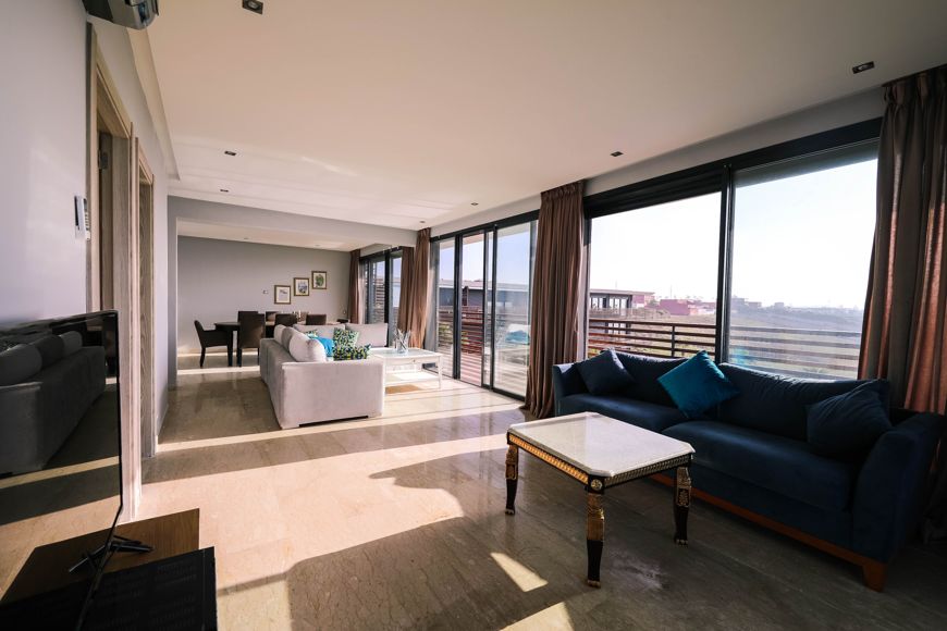 Spacious living room with large windows, beige tiled floor, blue and white sofas, a wooden coffee table, and views of a sunny balcony outside. Dining area is visible in the background.