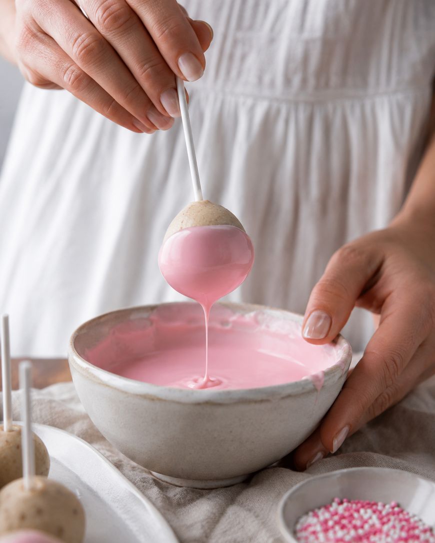 A person dips a cake pop into a bowl of pink icing. The cake pop is on a stick, and pink icing drips back into the bowl. The person wears a white dress, and a dish with pink and white sprinkles sits nearby.