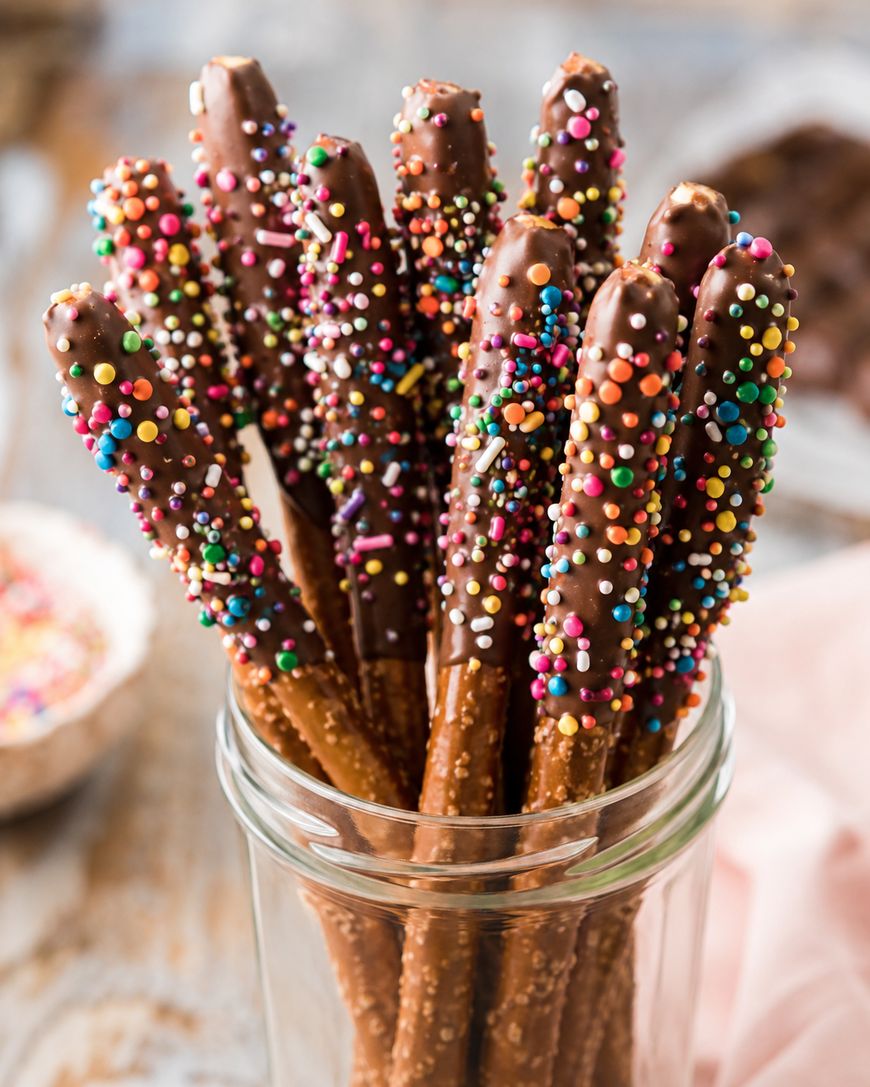 A glass jar filled with pretzel rods dipped in chocolate and coated with colorful sprinkles, standing upright on a wooden surface.