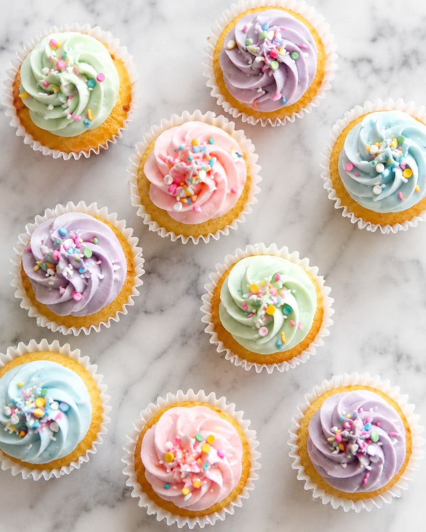 Ten cupcakes with pastel-colored swirled frosting and colorful sprinkles are arranged on a white marble surface. The cupcakes have frosting in pink, purple, blue, and green shades.