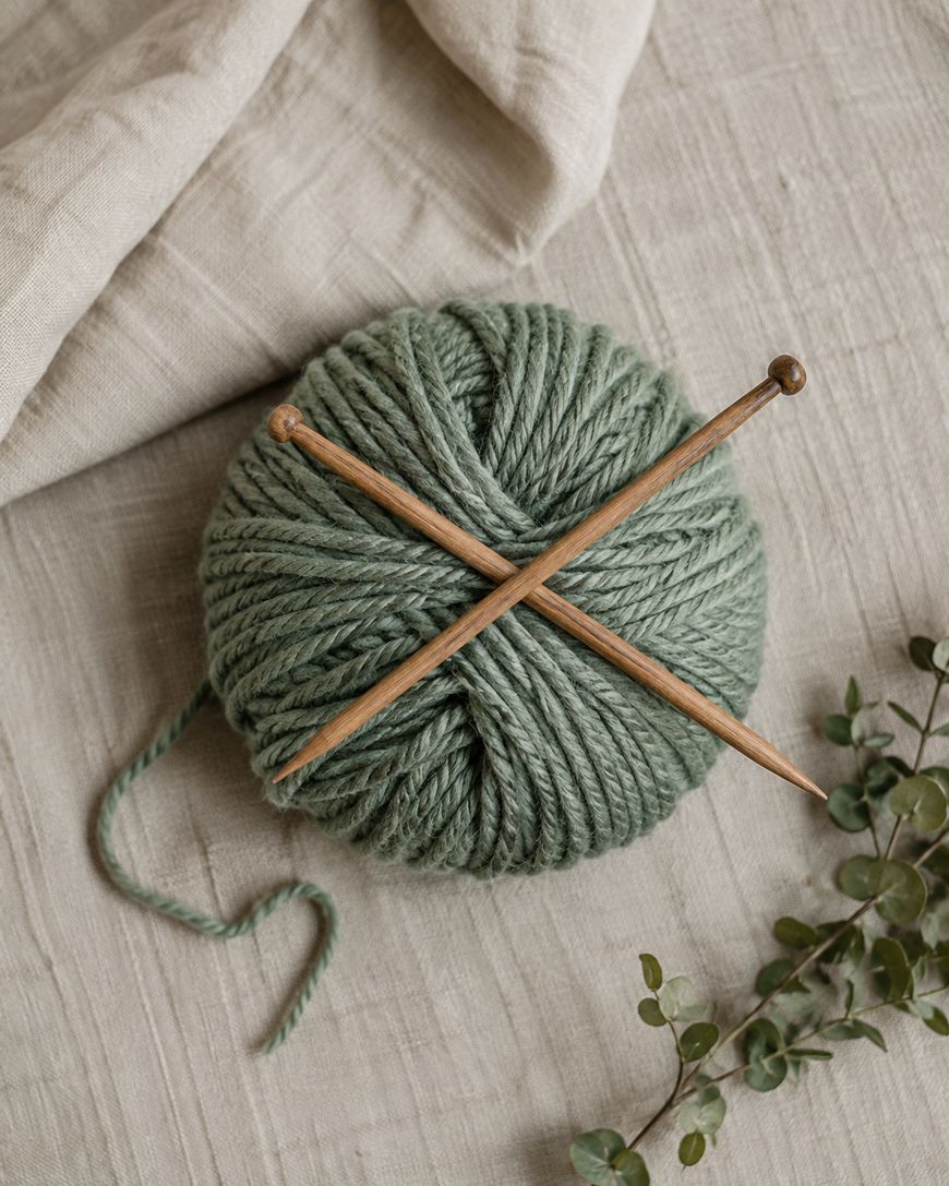 A green ball of yarn with two wooden knitting needles crossed on top, placed on a beige fabric surface with a small sprig of greenery nearby.