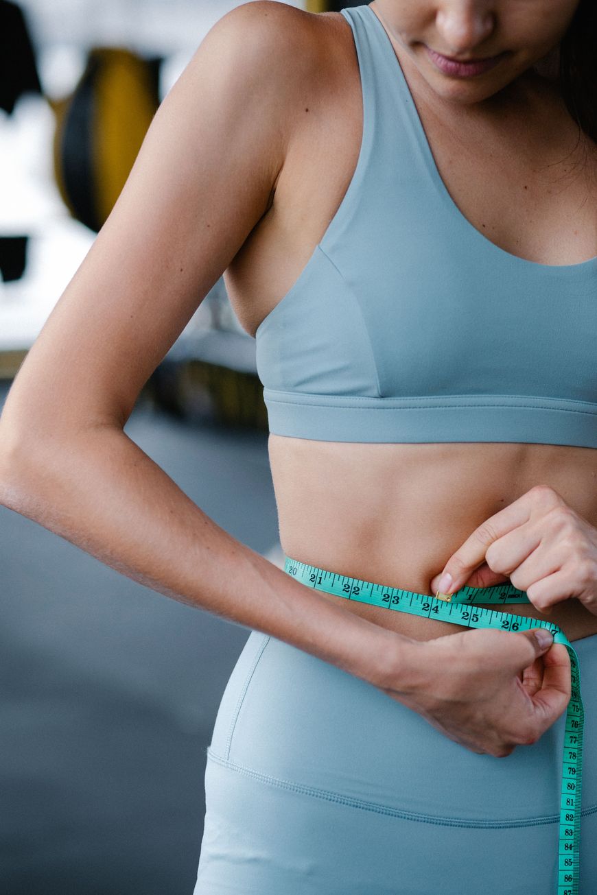 A woman in a light blue sports bra and leggings measures her waist with a blue measuring tape in a gym setting.