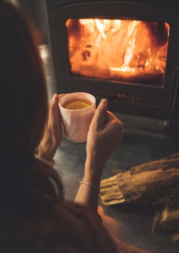 A person holding a white mug sits near a wood-burning stove with a warm fire, with logs placed beside them.