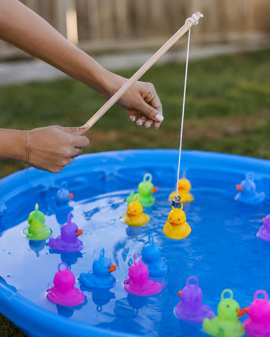 A person uses a wooden fishing pole to catch a yellow rubber duck from a blue kiddie pool filled with colorful rubber ducks on a grassy lawn.