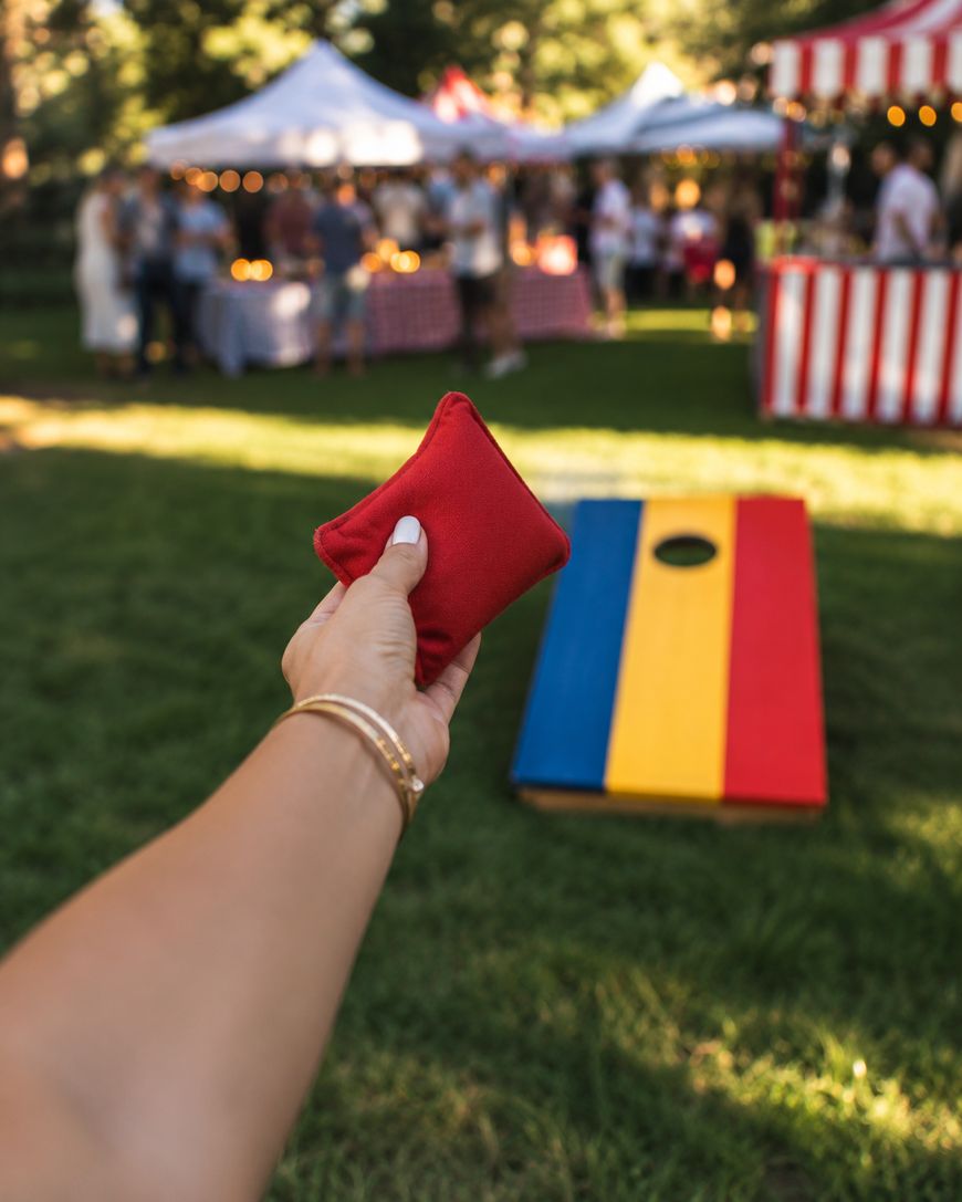 A person holding a red beanbag aims toward a colorful cornhole board at an outdoor event with tents and people in the background.