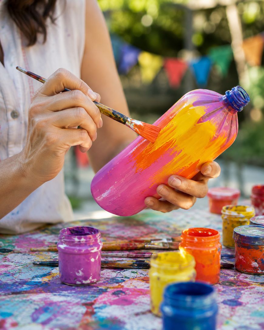 A person paints a pink plastic bottle with yellow and orange paint. Several colorful paint jars are on the table, and a festive banner hangs in the blurred background.