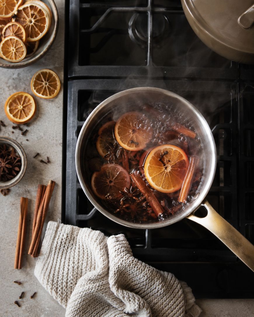 A pot on a stove simmers with sliced oranges, cinnamon sticks, and cloves. Steam rises, and dried orange slices, star anise, and a knitted towel are nearby on the counter.