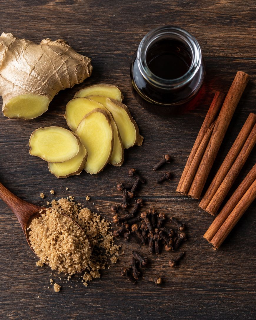 Sliced ginger, whole ginger root, cinnamon sticks, whole cloves, brown sugar on a wooden spoon, and a small glass jar of dark liquid arranged on a dark wooden surface.