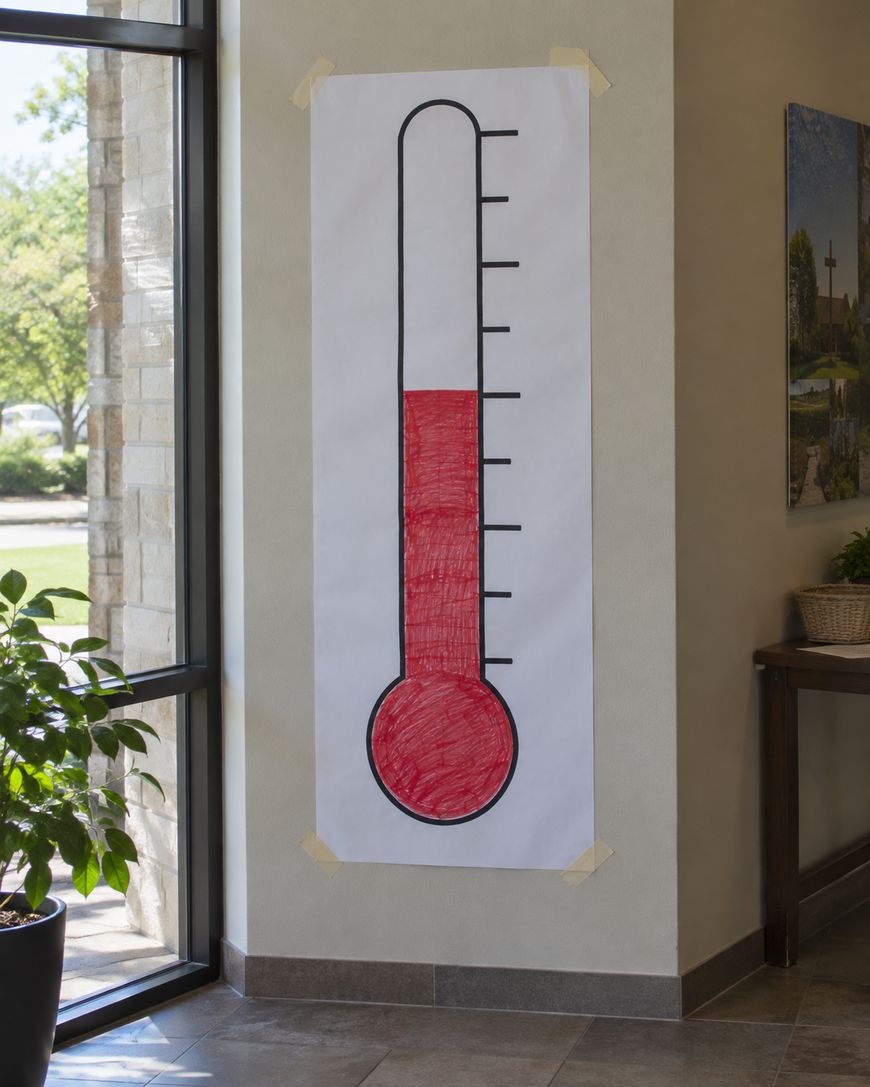 A large paper thermometer chart taped to a wall shows the level filled in red up to about halfway. The chart is near a window, with a green potted plant nearby.