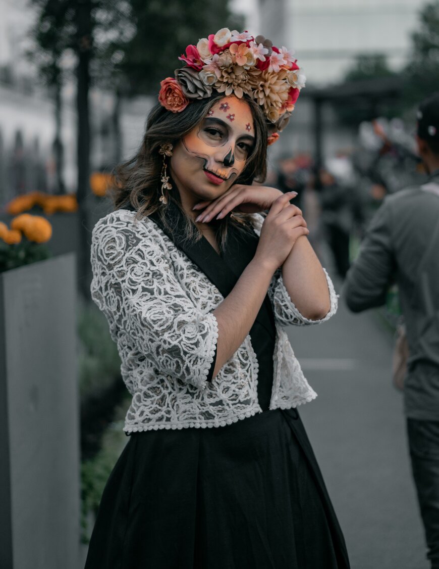 A woman with sugar-skull face makeup, a flower crown, and a white lace top for a Halloween costume.