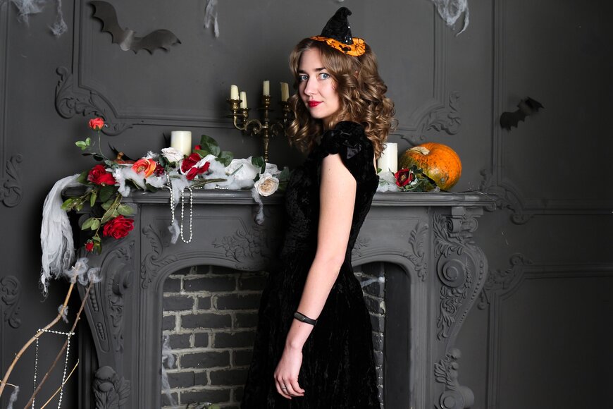 A woman in a black velvet dress and small witch hat fascinator beside a decorated mantel.