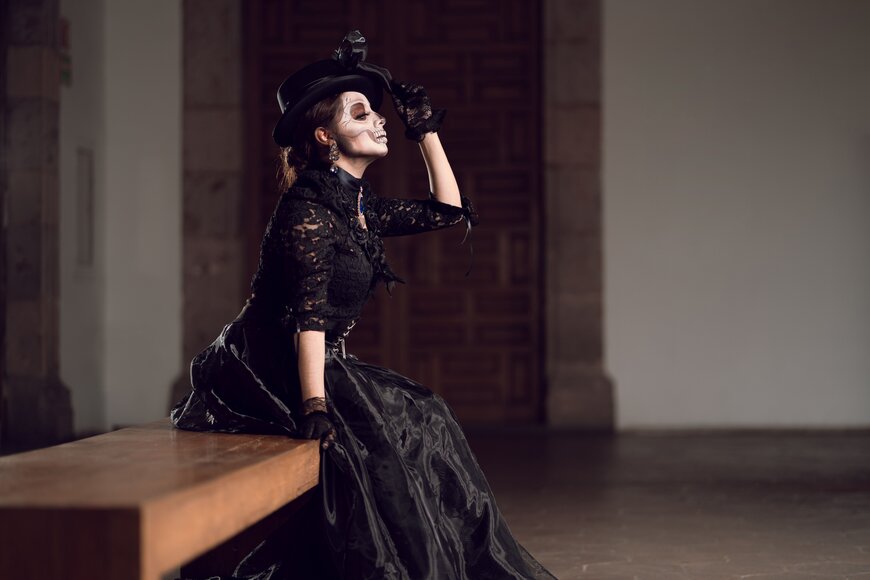 A woman in a Victorian black lace dress and top hat with skull-inspired makeup for a gothic Halloween costume.