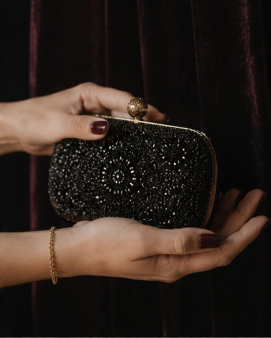 A woman's hands holding a small beaded clutch bag against a dark velvet backdrop.