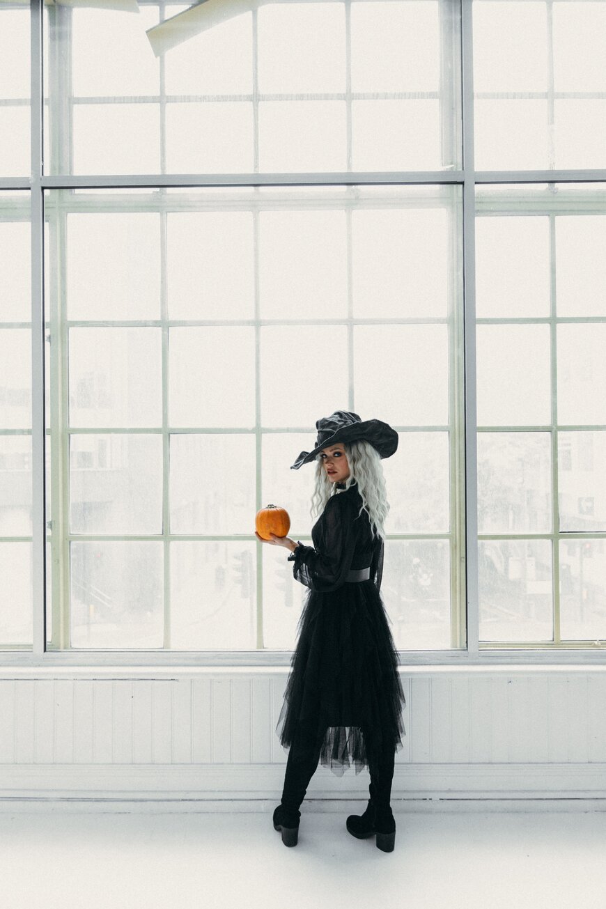 A woman in a wide-brim witch hat and black tulle dress holding a small pumpkin.