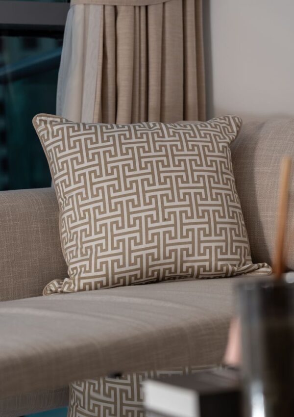 A beige sofa with a decorative pillow featuring a geometric Greek key pattern in white and tan. Neutral curtains and a window are in the background, with a blurred glass surface in the foreground.