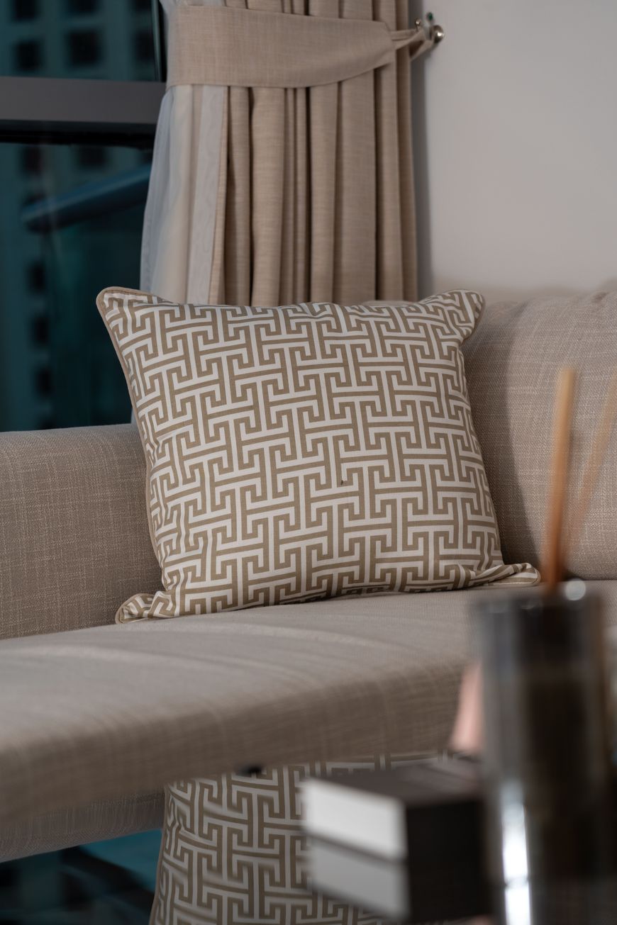 A beige sofa with a geometric-patterned cushion sits next to beige curtains. The scene is softly lit, and a glass table with blurred objects is in the foreground.
