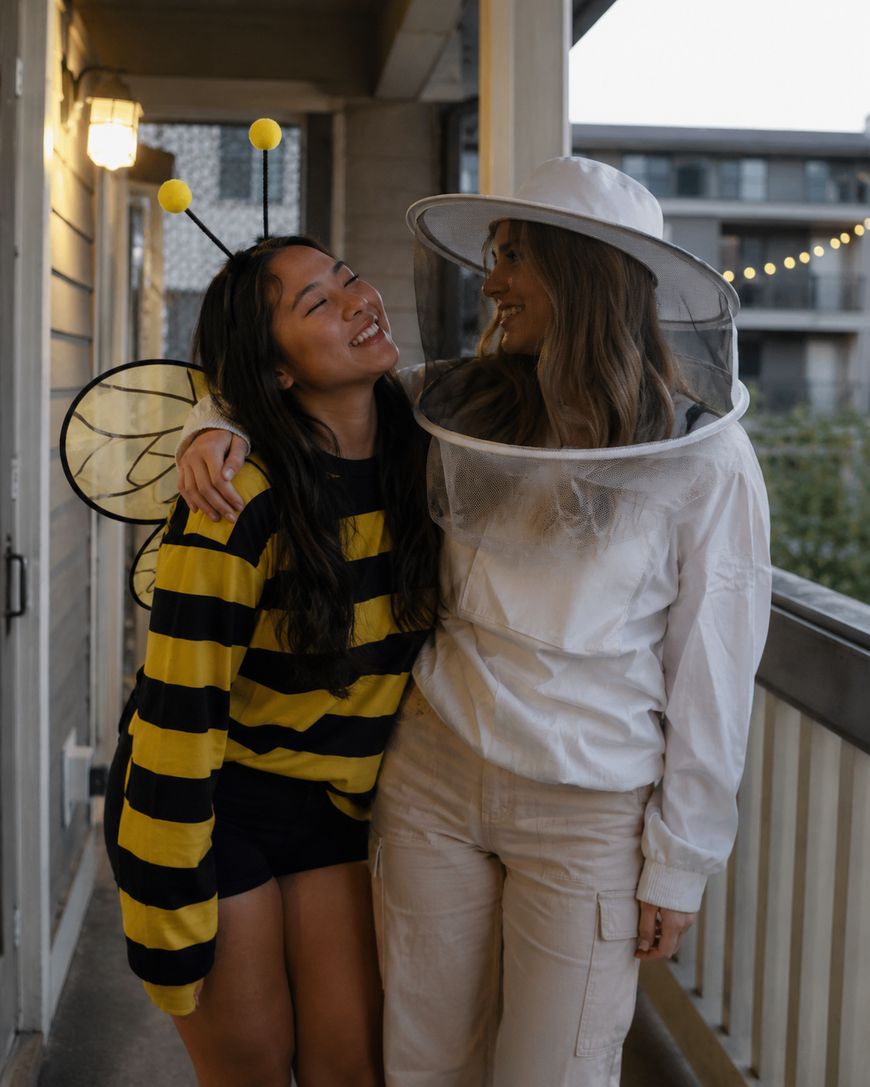 Two women in costumes smile at each other on a balcony. One wears a bee costume with wings and antennae, while the other is dressed as a beekeeper in white with a mesh veil hat.