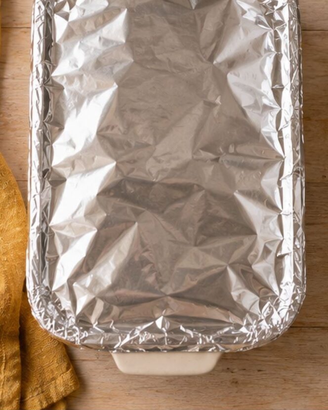 A rectangular baking dish covered with crinkled aluminum foil sits on a wooden surface next to a yellow cloth napkin.