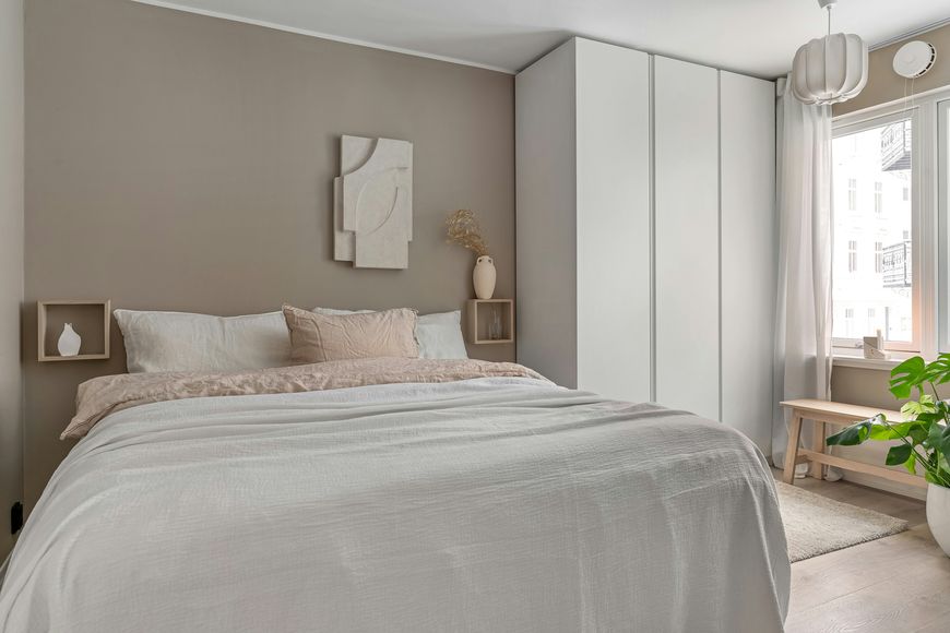 Minimalist bedroom with a beige accent wall, white bedding, built-in white wardrobe, abstract wall art, decorative vases on shelves, large window, small bench, and a potted plant by the window.