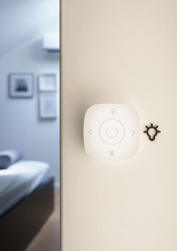 A smart wall switch with a power symbol and icons is mounted on a light-colored wall next to a small light bulb icon, with a blurred bedroom visible in the background.