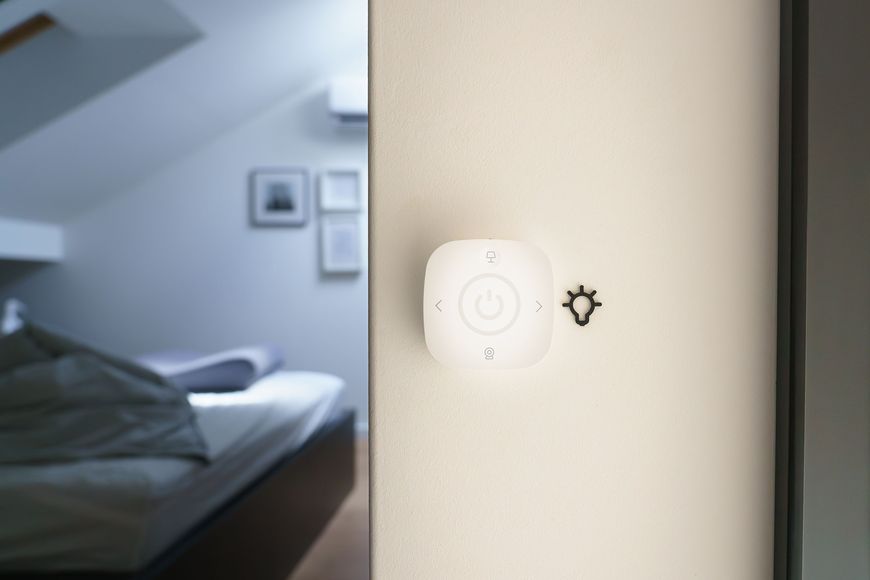 A smart light switch with touch controls is mounted on a white wall next to a symbol of a sun, with a blurred bedroom in the background featuring an unmade bed and framed pictures.