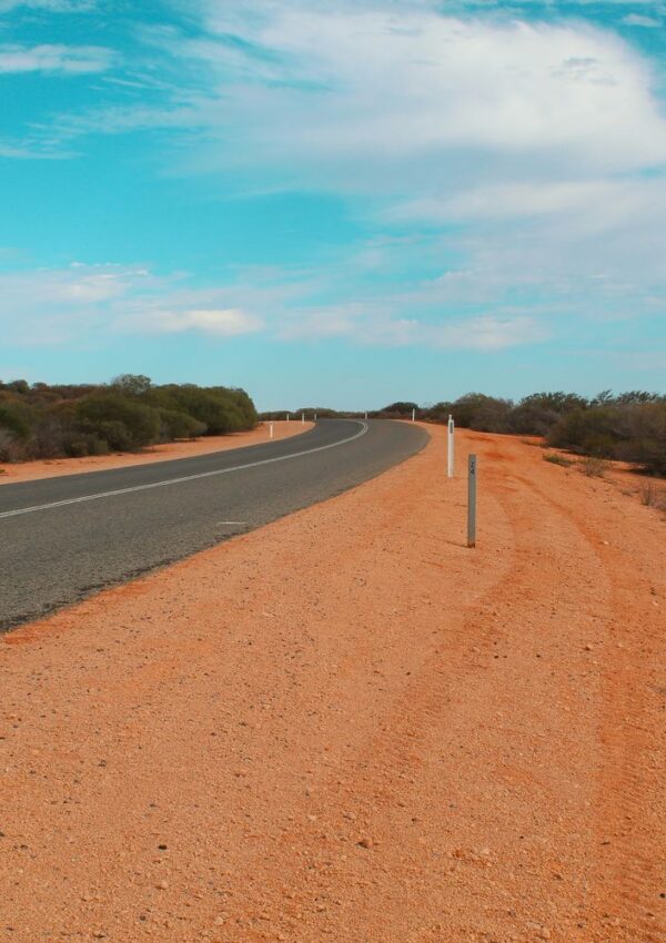 A paved road winds through a dry, reddish landscape with low bushes under a blue sky with scattered clouds, typical of the scenic drives near Kakadu. White road markers line the edge of the road, inviting travelers from Darwin to explore by car rental.