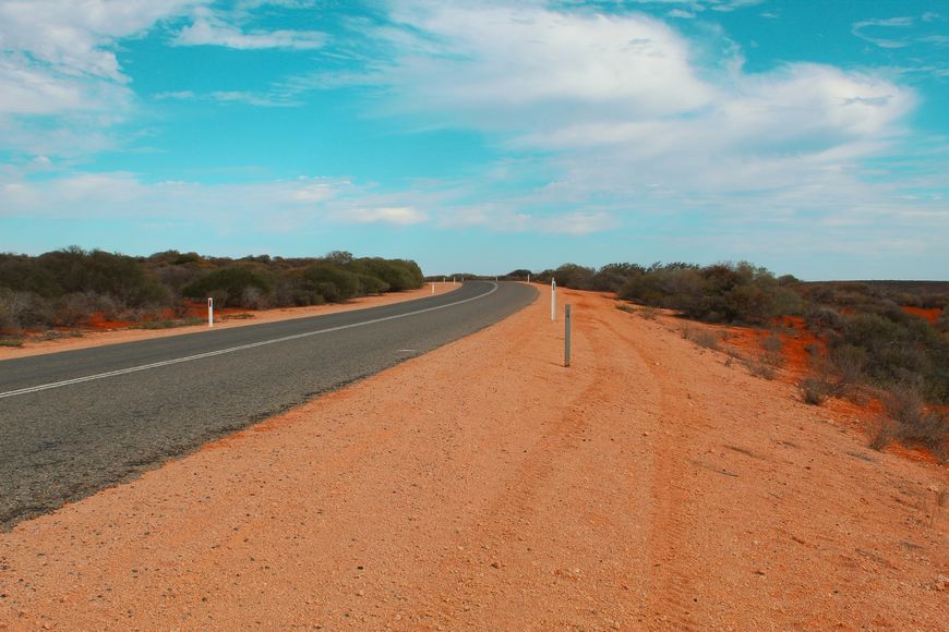 A paved road curves through a dry, desert landscape with sparse bushes on both sides under a partly cloudy blue sky, reminiscent of the scenic drives near Darwin or Kakadu—perfect for exploring by car rental.