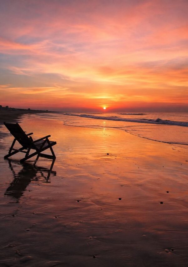 A wooden lounge chair sits on a sandy beach at sunset, with vibrant orange and pink clouds reflected in the wet sand and gentle waves in the background.
