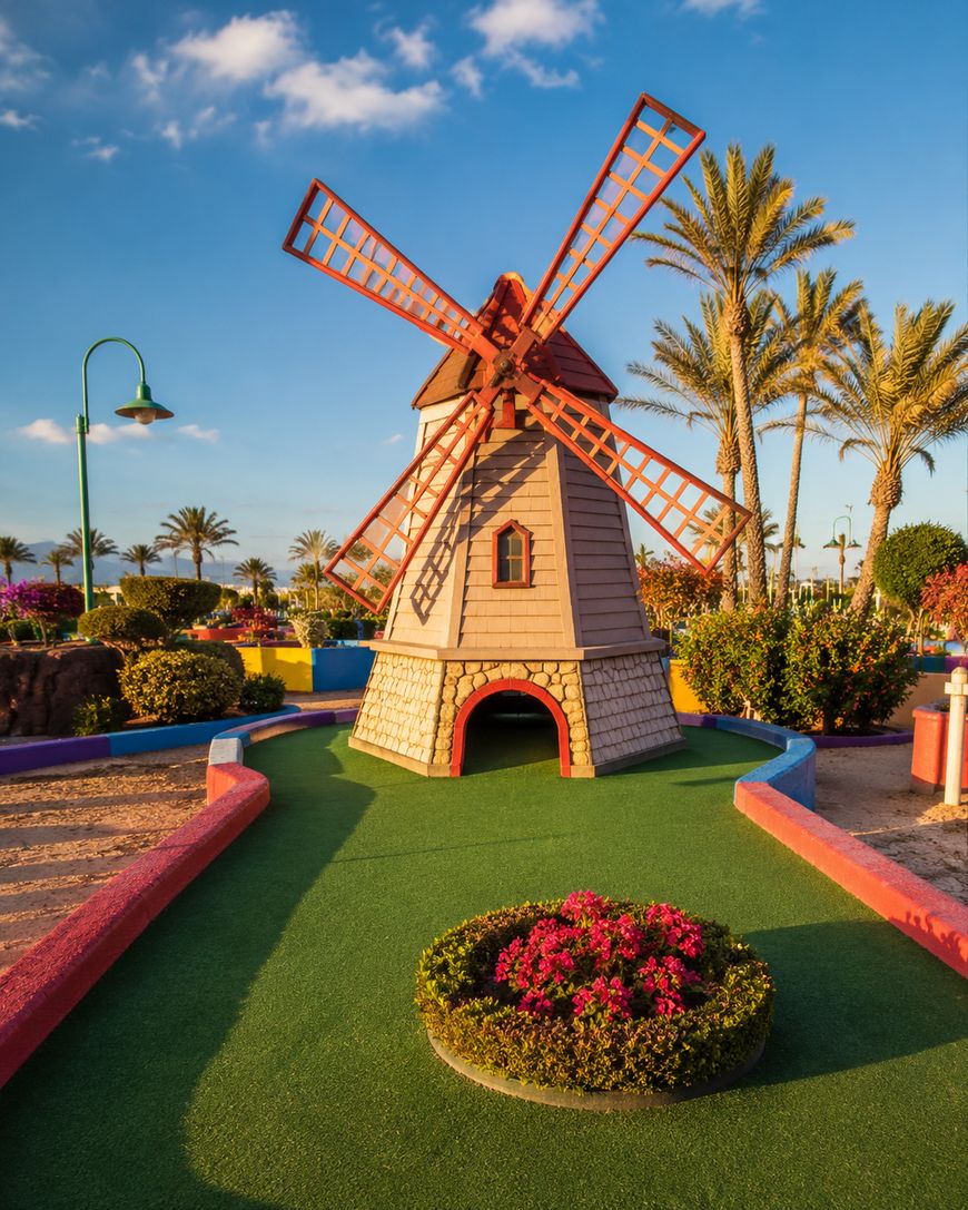A colorful mini-golf course with a windmill obstacle, surrounded by palm trees, vibrant flowers, and a bright blue sky.